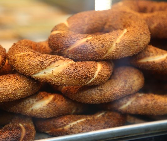 Simit, Osmanlı döneminden günümüze kadar gelen köklü bir geçmişe sahiptir. İlk olarak saray mutfaklarında farklı un karışımlarıyla yapılan bu lezzet, zamanla halk arasında yaygınlaşarak günlük yaşamın bir parçası haline gelmiştir. Özellikle 16. yüzyıldan itibaren İstanbul’da simit fırınlarının açılmasıyla birlikte, bu eşsiz yiyecek şehir kültürünün önemli bir unsuru olmuştur. Bugün Anadolu’nun dört bir yanında farklı isimlerle anılsa da, ortak nokta simidin sofralarda her daim yer bulmasıdır. Konulu bir haber görseli.