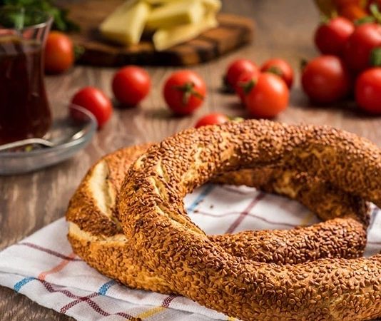 Türkiye’de sokak lezzetleri denildiğinde akla gelen ilk tatlar arasında simit ve açma yer alır. Günün her saatinde ulaşılabilen bu iki hamur işi, yalnızca pratik bir atıştırmalık olmanın ötesinde, şehir yaşamının ritmini belirleyen önemli unsurlardan biri olarak görülür. Sabah işe yetişmeye çalışan bir memurun elinde, okul yolunda koşan bir öğrencinin çantasında ya da sahil kenarında çay eşliğinde tüketilen bu lezzetler, toplumsal hafızanın da güçlü bir parçasıdır. Konulu bir haber görseli.